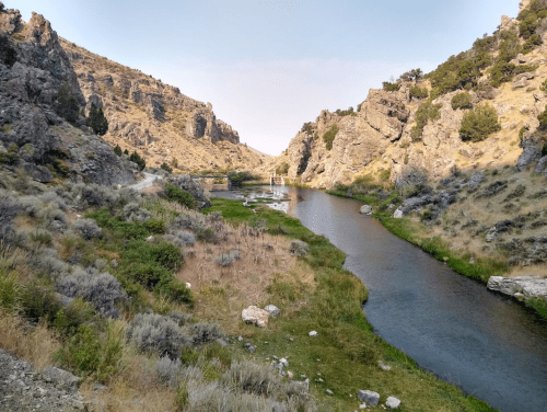 A serene river flows through a rocky canyon, surrounded by lush greenery and rugged hills under a clear sky.
