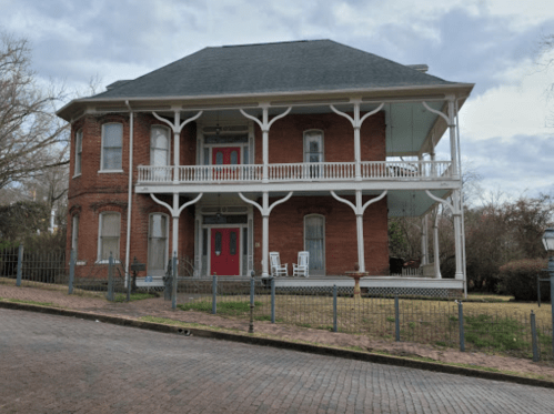 A two-story brick house with a gray roof, red doors, and a porch, surrounded by a fence and grassy area.