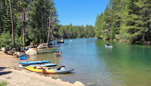 A serene lake surrounded by trees, with people kayaking and swimming, and colorful kayaks lined up on the shore.