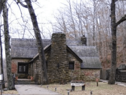 A rustic stone house surrounded by bare trees, with a pathway leading to a sign that reads "Spring House."