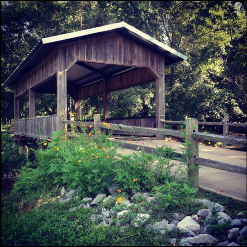 A wooden covered bridge surrounded by greenery and colorful flowers, with a path leading through it.