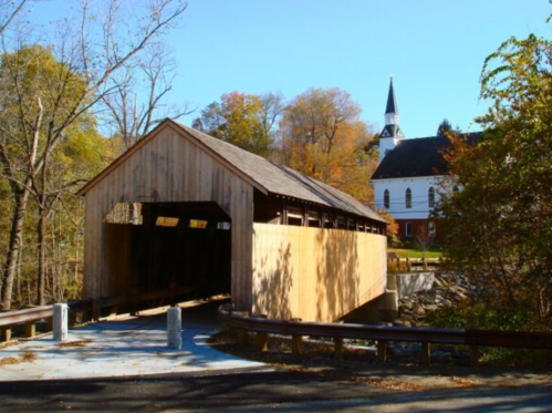 A wooden covered bridge spans a small river, with a church and autumn trees in the background.