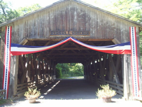 A wooden covered bridge with patriotic red, white, and blue decorations at the entrance, surrounded by greenery.