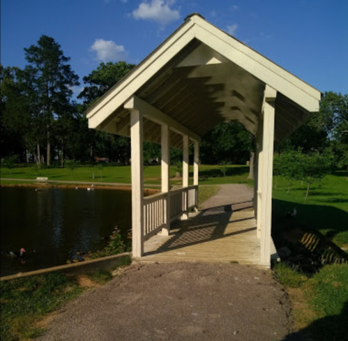 A wooden gazebo by a pond, surrounded by green grass and trees, with ducks visible in the water.
