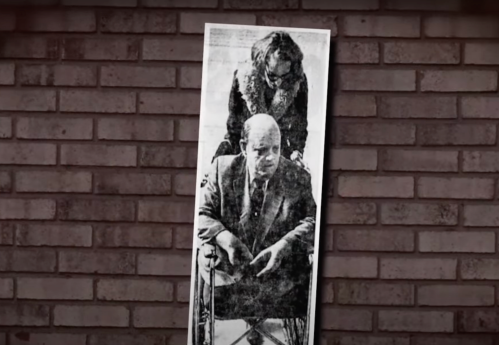 A black-and-white photo of a man in a wheelchair, with a woman standing behind him, against a brick wall background.