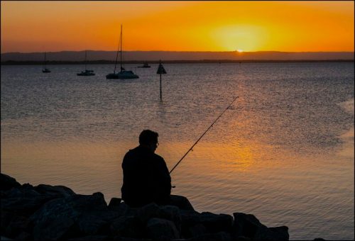 A person fishing on a rocky shore at sunset, with boats in the water and a vibrant orange sky.
