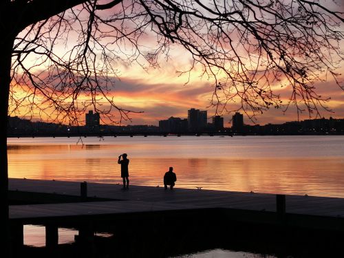 Silhouettes of two people by a lake at sunset, with colorful clouds and city skyline in the background.