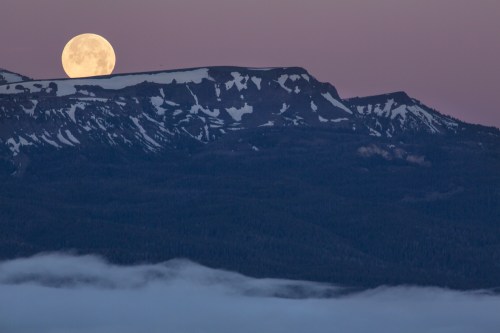 A full moon rises over snow-capped mountains, casting a serene glow against a twilight sky.