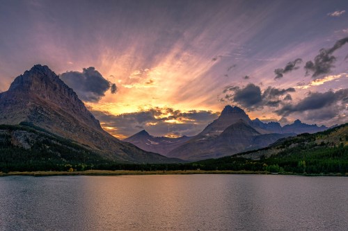 A serene lake reflects a vibrant sunset behind majestic mountains and a colorful sky.