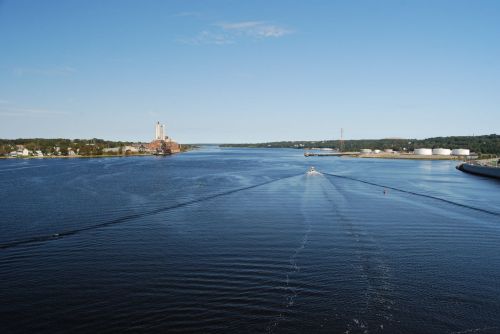 A serene river scene with a boat leaving a wake, flanked by trees and industrial buildings under a clear blue sky.
