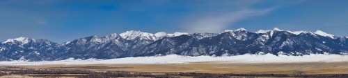 Panoramic view of snow-capped mountains under a clear blue sky, with a grassy plain in the foreground.