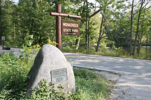 Sign for Monadnock State Park next to a large rock, surrounded by trees and a road in a natural setting.