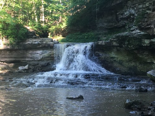 A serene waterfall cascading over rocky ledges into a calm pool, surrounded by lush greenery and natural rock formations.