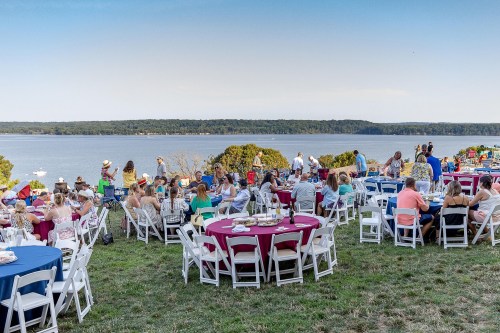A lively outdoor event by the water, with guests seated at colorful tables enjoying the scenic view.