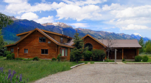 A rustic wooden house surrounded by greenery, with mountains in the background under a partly cloudy sky.