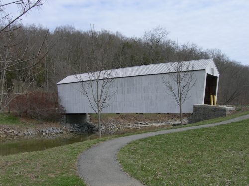 A covered bridge stands over a stream, surrounded by bare trees and a grassy path leading to it.