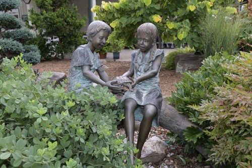 Two bronze statues of girls sitting together, sharing a small object, surrounded by lush greenery and rocks.