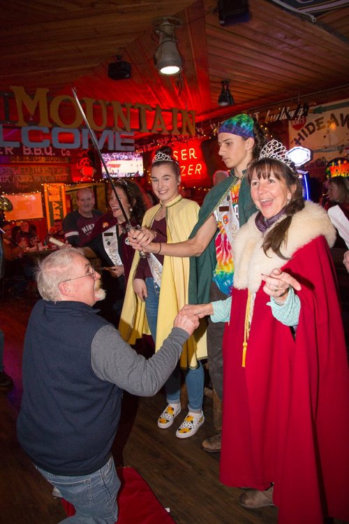 A festive scene in a restaurant with people in costumes, including tiaras, engaging in a playful sword ceremony.