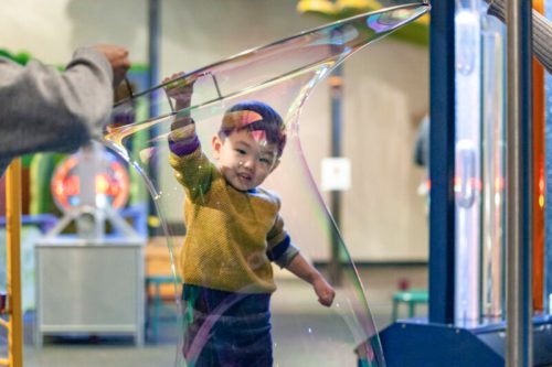 A child smiles while being surrounded by a large soap bubble at a playful indoor exhibit.