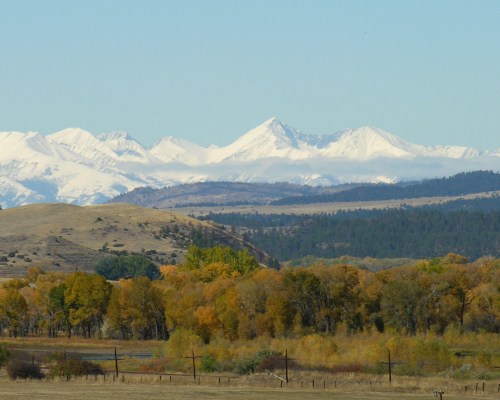 Snow-capped mountains rise above a landscape of autumn trees and rolling hills under a clear blue sky.