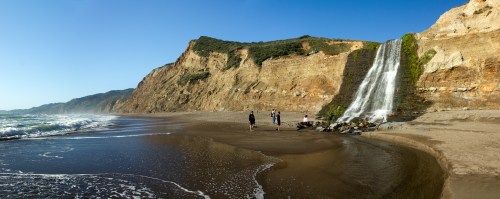 A scenic beach with a waterfall cascading down a cliff, and three people walking along the sandy shore.