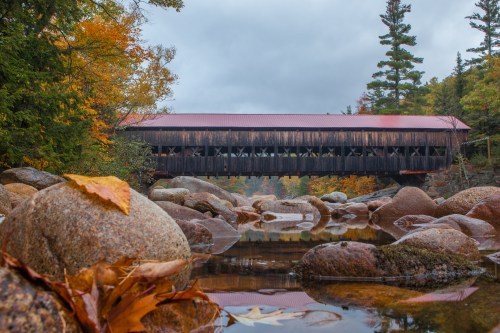 A covered bridge spans a rocky river, surrounded by autumn foliage and overcast skies.