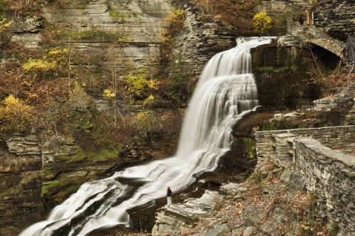 A serene waterfall cascades over rocky cliffs, surrounded by autumn foliage and a person standing nearby.