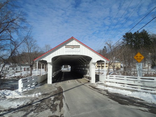 A snowy scene featuring a covered bridge with a red roof and white walls, surrounded by trees and a clear blue sky.