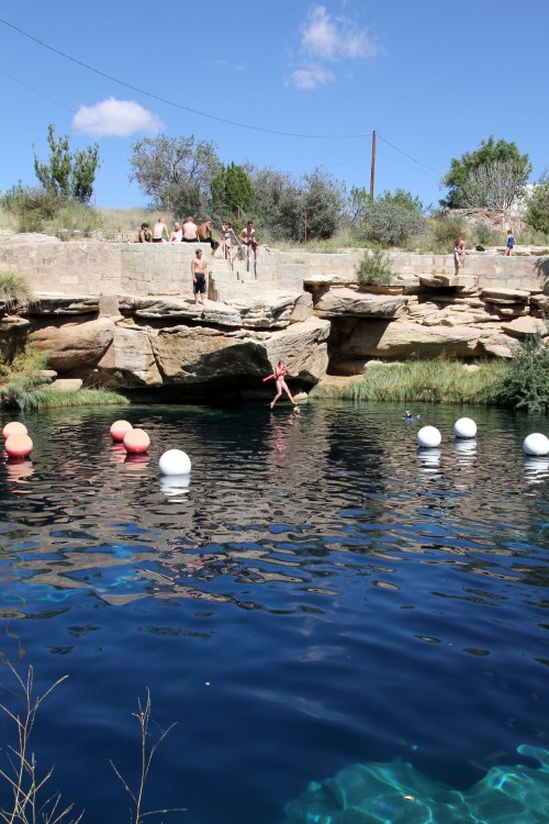 A natural swimming hole with people jumping in, surrounded by rocks and greenery under a clear blue sky.
