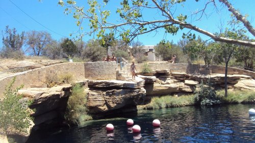 A natural swimming area with rocky edges, people relaxing on the shore, and colorful buoys in the water under a clear sky.