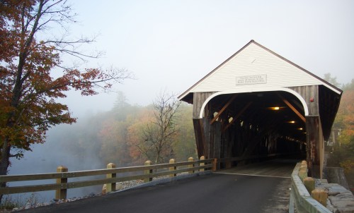 A covered bridge surrounded by fog and autumn foliage, with a road leading into the structure.