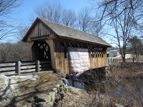 A wooden covered bridge with an American flag draped on the side, surrounded by bare trees and a calm stream.