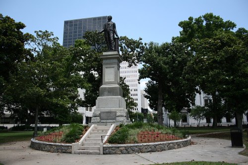 A statue on a pedestal surrounded by greenery in a park, with buildings visible in the background.