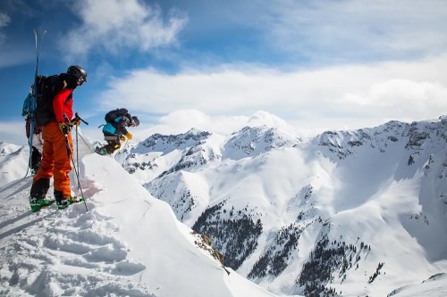 Two skiers on a snowy mountain ridge, one preparing to jump off a ledge, with majestic peaks in the background.
