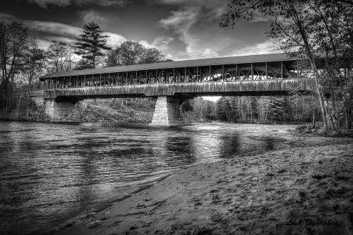 A black and white image of a covered bridge spanning a river, surrounded by trees and a sandy riverbank.