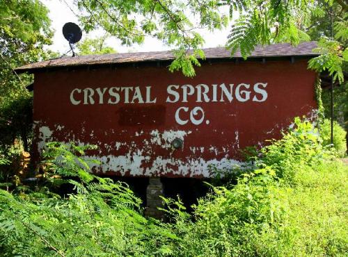 A weathered red building with "CRYSTAL SPRINGS CO." painted on it, surrounded by overgrown greenery.