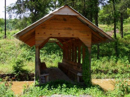 A wooden shelter with a slanted roof, surrounded by greenery and a small stream flowing nearby.