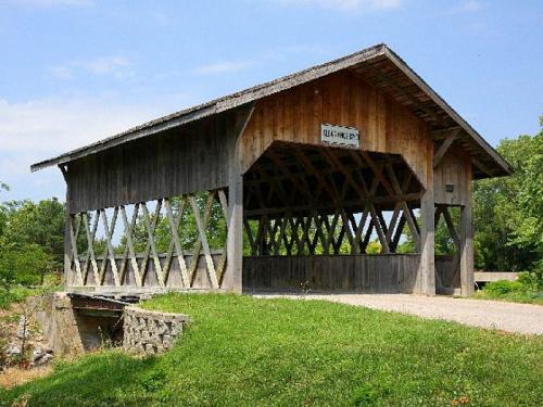 A wooden covered bridge with a peaked roof, set against a blue sky and green landscape.
