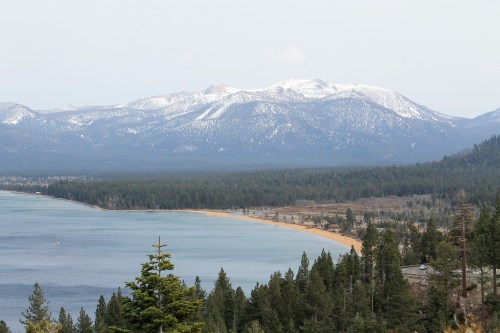 Scenic view of a lake bordered by a sandy beach and forest, with snow-capped mountains in the background.