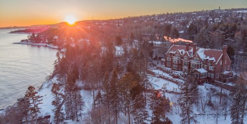 A scenic winter sunset over a lake, highlighting a large house surrounded by snow-covered trees.