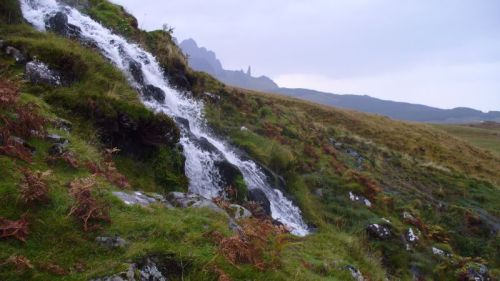 A small waterfall cascades down rocky terrain, surrounded by lush green grass and distant mountains under a cloudy sky.