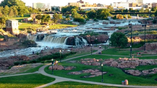 A scenic view of a waterfall cascading over rocks in a park, surrounded by greenery and city buildings in the background.