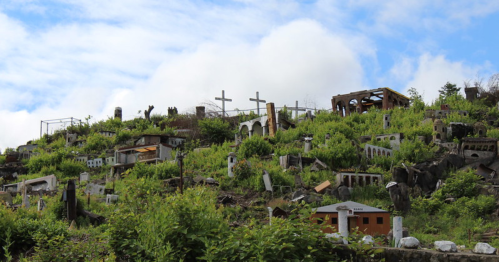 A hillside cemetery overgrown with greenery, featuring various tombstones and crosses under a cloudy sky.