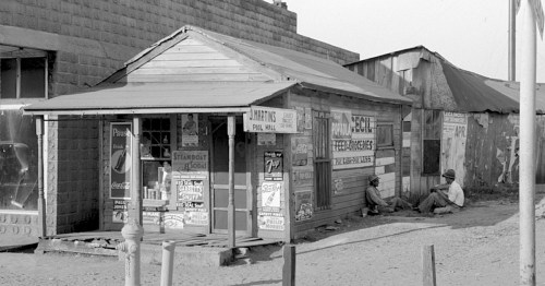 Black and white image of a rustic storefront with vintage advertisements, two men sitting nearby on the ground.