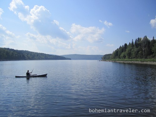 A person kayaking on a calm lake surrounded by lush green trees and a clear blue sky.