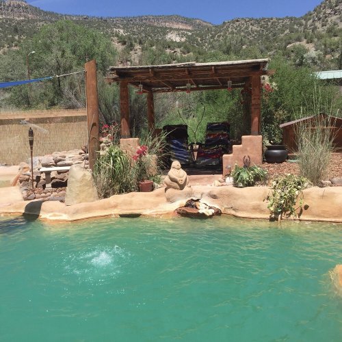 A serene outdoor scene featuring a turquoise pool, a shaded seating area, and lush greenery in the background.