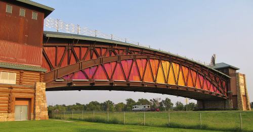 A colorful arch bridge spans over a grassy area, with a rustic building on one side and a camper in the background.