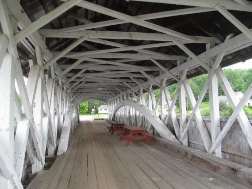 Interior view of a covered bridge featuring white wooden beams and picnic tables along the walkway.