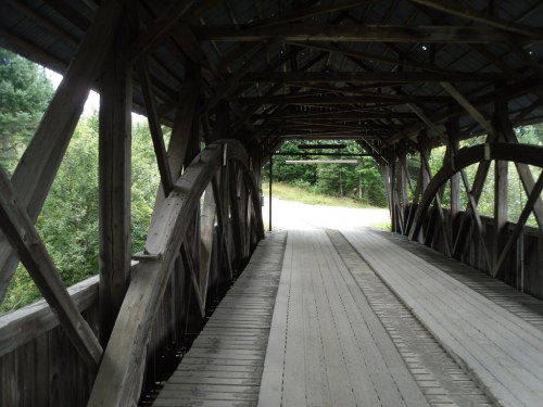 Interior view of a wooden covered bridge with a rustic design, leading to a grassy path surrounded by trees.