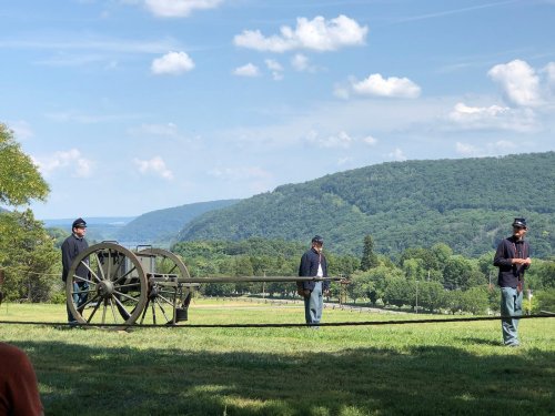 Three historical reenactors in Civil War uniforms operate a cannon on a grassy field with rolling hills in the background.